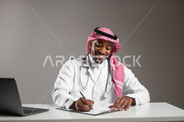 اجراء الاستشارات الطبيه عن بعد، استخدام التكنولوجيا في البحث والتطوير، تقديم الخدمات للمرضى بكل كفاءه، بورتريه مقرب لطبيب عربي خليجي سعودي يرتدي الثوب التقليدي والمعطف الابيض يقوم بتدوين وتسجيل البيانات، ايماءات السرور والسعادة، مهن ووظائف صحية، خلفيه رماديه