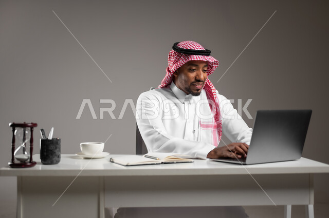 استخدام الأجهزة والتقنيات الحديثة والمتطورة، عقد اجتماع عمل عبر الانترنت، بورتريه مقرب لرجل عربي خليجي سعودي مبتسم يرتدي الثوب التقليدي والشماغ يعمل على جهاز الحاسوب المحمول، تناول المشروبات الساخنة، العمل في المجال المكتبي، خلفية رمادية