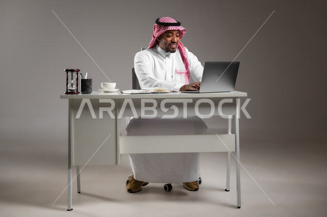 العمل في المجال المكتبي، عقد اجتماع عمل عبر الانترنت، استخدام الأجهزة والتقنيات الحديثة والمتطورة، بورتريه لرجل عربي خليجي سعودي يرتدي الثوب التقليدي والشماغ يعمل على جهاز الحاسوب المحمول، تناول المشروبات الساخنة، صورة لكامل الجسم، خلفية رمادية