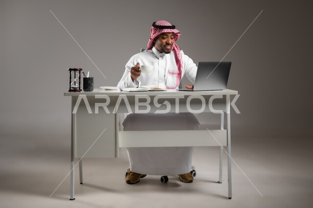 Browsing social media on a computer, holding a business meeting over the Internet, using modern and advanced devices and technologies, portrait of a Saudi Arabian Gulf man wearing a traditional thobe and shemagh holding a cup of coffee, working in the office, full body image, gray background