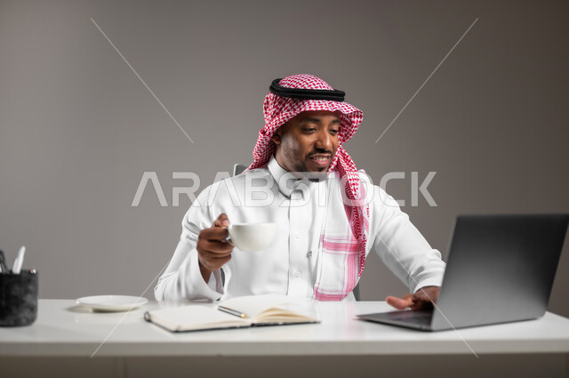 Using modern and advanced devices and technologies, holding a business meeting via the Internet, close-up portrait of a smiling Saudi Arabian Gulf man wearing traditional thobe and shemagh holding a cup of coffee, browsing social media sites on a computer, working in the office field, gray background