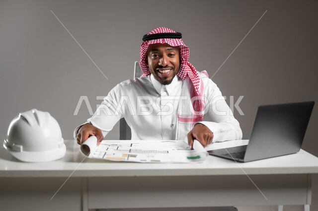 تدقيق ومراجعة المخططات الإنشائية، العمل في القطاع الهندسي، إدارة المشاريع الهندسية، ايماءات السرور والسعادة، دمج التكنولوجيا بالعمل، بورتريه مقرب لمهندس عربي خليجي سعودي يرتدي الثوب التقليدي يجلس على المكتب يقوم بدراسة أساسيات المشروع، خلفية رمادية