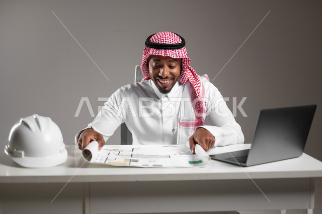 إدارة المشاريع الهندسية، ايماءات السرور والسعادة، دمج التكنولوجيا بالعمل، دراسة أساسيات المشروع، العمل في القطاع الهندسي، بورتريه مقرب لمهندس عربي خليجي سعودي يرتدي الثوب التقليدي يجلس على المكتب يقوم بتدقيق ومراجعة المخططات الإنشائية، خلفية رمادية
