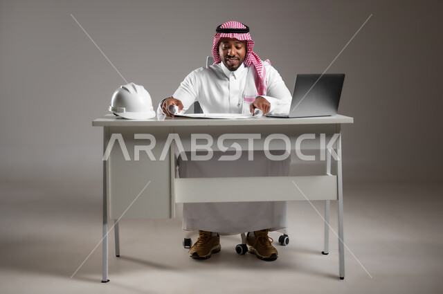 Engineering project management, integrating technology into work, studying project basics, working in engineering sector, portrait of a Saudi Arabian Gulf engineer wearing traditional thobe and safety vest sitting at desk checking and reviewing construction plans, full body image, gray background