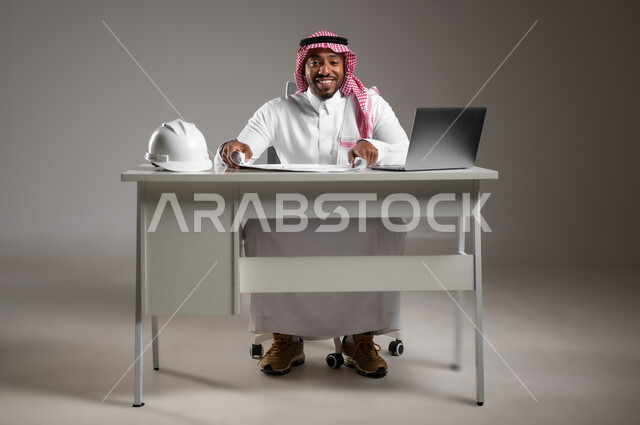 Working in engineering sector, studying project basics, engineering project management, gestures of pleasure and happiness, integrating technology into work, portrait of a Saudi Arabian Gulf engineer wearing traditional dress sitting at desk checking and reviewing construction plans, full body image, gray background