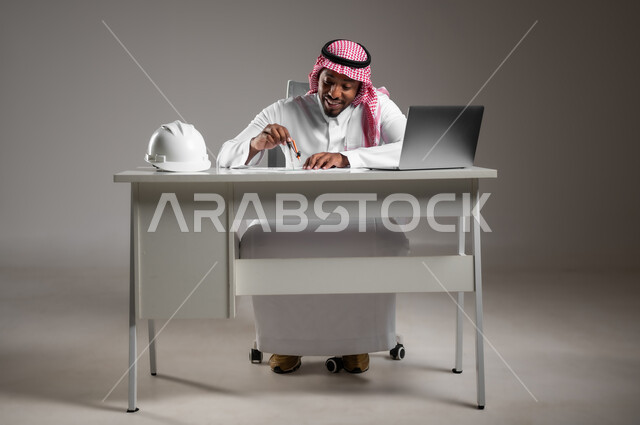 العمل في القطاع الهندسي، دراسة أساسيات المشروع، إدارة المشاريع الهندسية، ايماءات السرور والسعادة، دمج التكنولوجيا بالعمل، بورتريه لمهندس عربي خليجي سعودي يرتدي الثوب التقليدي يجلس على المكتب يقوم بتدقيق ومراجعة المخططات الإنشائية، صورة لكامل الجسم، خلفية رمادية