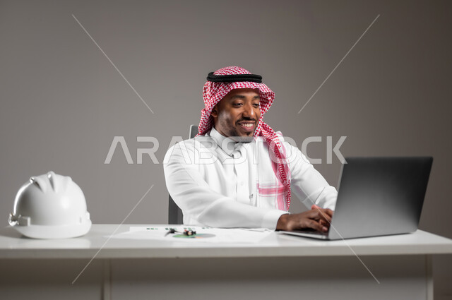 استخدام الأجهزة التقينة الحديثة، دراسة أساسيات المشروع، تدقيق ومراجعة المخططات الإنشائية، العمل في القطاع الهندسي، دمج التكنولوجيا بالعمل، بورتريه مقرب لمهندس عربي خليجي سعودي يرتدي الثوب التقليدي يجلس على المكتب يعمل على الحاسوب المحمول، خلفية رمادية