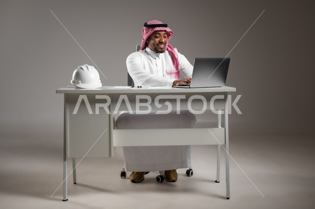 دمج التكنولوجيا بالعمل، دراسة أساسيات المشروع، تدقيق ومراجعة المخططات الإنشائية، العمل في القطاع الهندسي، بورتريه لمهندس عربي خليجي سعودي يرتدي الثوب التقليدي والشماغ يجلس على المكتب يعمل على الحاسوب المحمول، صورة لكامل الجسم، خلفية رمادية