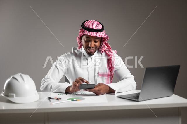 استخدام الأجهزة التقينة الحديثة، دراسة أساسيات المشروع، تدقيق ومراجعة المخططات الإنشائية، العمل في القطاع الهندسي، دمج التكنولوجيا بالعمل، بورتريه مقرب لمهندس عربي خليجي سعودي يرتدي الثوب التقليدي يجلس على المكتب يعمل على الحاسوب المحمول، خلفية رمادية