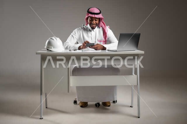 Integrating technology into work, studying project basics, auditing and reviewing construction plans, working in engineering sector, portrait of a Saudi Arabian Gulf engineer wearing traditional thobe and shemagh sitting at desk working on laptop, full body photo, gray background