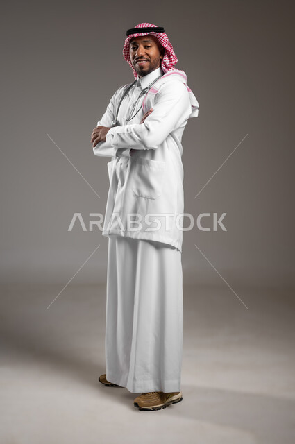 النظر الكاميرا بايماءات الثقة بالنفس، العمل في القطاع الصحي، مفهوم الفحص الطبي، تقديم الخدمات العلاجية للمرضى, بورتريه من الجانب لطبيب عربي خليجي سعودي يرتدي الزي التقليدي والمعطف الطبي يضع السماعة الطبية يقف بأيدي متقاطعة، صورة لكامل الجسم، خلفية رمادية
