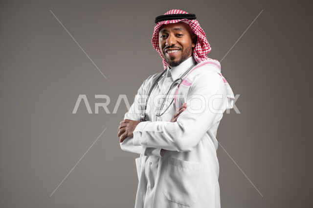 العمل في القطاع الصحي، النظر الكاميرا بايماءات الثقة بالنفس، مفهوم الفحص الطبي، تقديم الخدمات العلاجية للمرضى, بورتريه مقرب من الجانب لطبيب عربي خليجي سعودي يرتدي الزي التقليدي والمعطف الطبي يضع السماعة الطبية يقف بأيدي متقاطعة، خلفية رمادية
