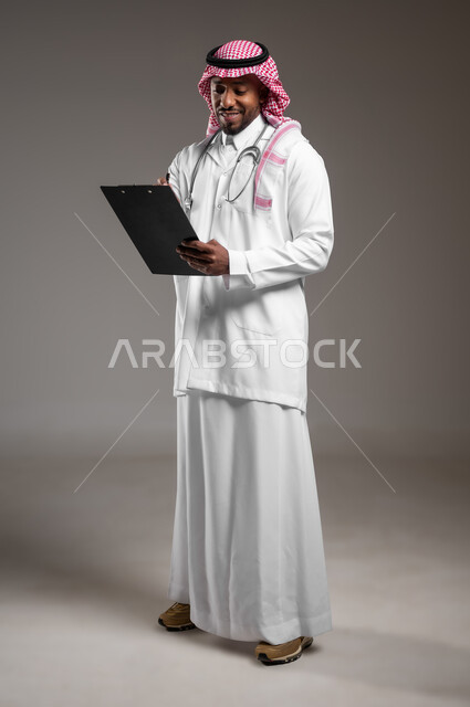العمل في القطاع الصحي، اعداد التقارير الطبية، تدوين نتائج الفحوصات بالسجل الطبي، حسن التعامل مع المراجعين، ايماءات البشاشة والسرور، بورتريه لطبيب عربي خليجي سعودي يرتدي الثوب التقليدي والمعطف الابيض يحمل بيده ملفات المرضى، صورة لكامل الجسم، خلفية رمادية