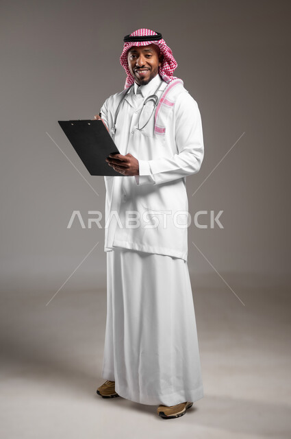اعداد التقارير الطبية، تدوين نتائج الفحوصات بالسجل الطبي، حسن التعامل مع المراجعين، ايماءات البشاشة والسرور، بورتريه لطبيب عربي خليجي سعودي يرتدي الثوب التقليدي والمعطف الابيض يحمل بيده ملفات المرضى، العمل في القطاع الصحي، صورة لكامل الجسم، خلفية رمادية