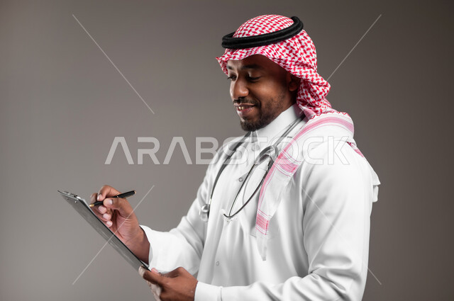 تدوين نتائج الفحوصات بالسجل الطبي، حسن التعامل مع المراجعين، ايماءات البشاشة والسرور، بورتريه مقرب من الجانب لطبيب عربي خليجي سعودي يرتدي الثوب التقليدي والمعطف الابيض يحمل بيده ملفات المرضى، اعداد التقارير الطبية، العمل في القطاع الصحي، خلفية رمادية