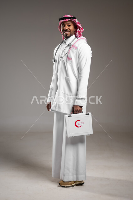 Providing emergency health services to patients, principles of dealing with emergency cases, side portrait of a Saudi Arabian Gulf doctor wearing traditional dress and coat holding a first aid kit, necessary medical tools and equipment, working in the health sector, full body image, gray background
