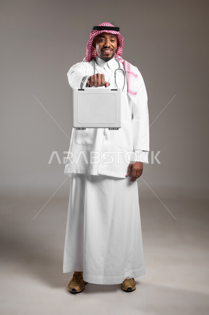 مبادئ التعامل مع حالات الطوارئ، تقديم الخدمات الصحية الطارئة للمرضى، بورتريه لطبيب عربي خليجي سعودي يرتدي الزي التقليدي والمعطف الابيض يحمل بيده حقيبة الاسعافات الأولية، الأدوات والمعدات الطبية اللازمة، العمل في القطاع الصحي، صورة لكامل الجسم، خلفية رمادية