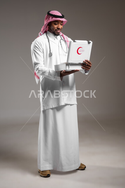 الأدوات والمعدات الطبية اللازمة، تقديم الخدمات الصحية الطارئة للمرضى، مبادئ التعامل مع حالات الطوارئ، بورتريه لطبيب عربي خليجي سعودي يرتدي الزي التقليدي والمعطف يحمل بيده حقيبة الاسعافات الأولية المفتوحة، العمل في القطاع الصحي، صورة لكامل الجسم، خلفية رمادية