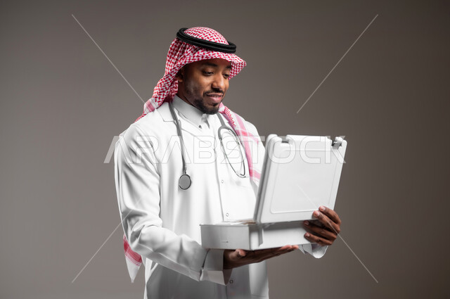 العمل في القطاع الصحي، الأدوات والمعدات الطبية اللازمة، تقديم الخدمات الصحية الطارئة للمرضى، مبادئ التعامل مع حالات الطوارئ، بورتريه مقرب لطبيب عربي خليجي سعودي يرتدي الزي التقليدي والمعطف يحمل بيده حقيبة الاسعافات الأولية المفتوحة، خلفية رمادية