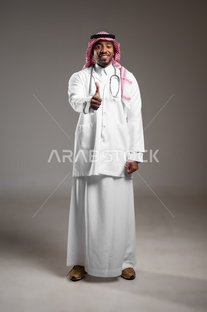مفهوم الفحص الطبي، ايماءات القبول والموافقة، العمل في القطاع الصحي، تقديم الخدمات العلاجية للمرضى, بورتريه لطبيب عربي خليجي سعودي يرتدي الزي التقليدي والمعطف الطبي يضع السماعة الطبية يرفع يده بتعابير الاعجاب والرضى، صورة لكامل الجسم، خلفية رمادية