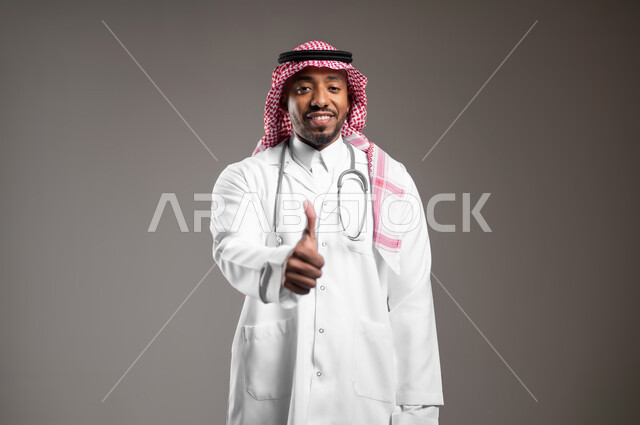 Working in the health sector, the concept of medical examination, providing medical services to patients, close-up portrait of a Saudi Arabian Gulf doctor wearing traditional clothing and a medical coat, wearing a stethoscope, raising his hand with expressions of admiration and satisfaction, gray background