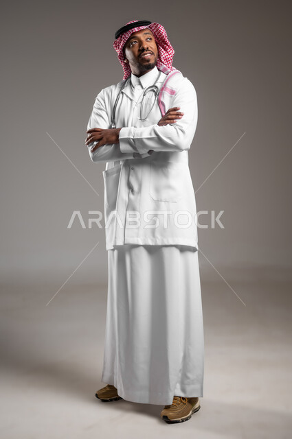 النظر لمكان ما بايماءات الثقة بالنفس، العمل في القطاع الصحي، الفحص الطبي وتقديم الخدمات العلاجية للمرضى, بورتريه لطبيب عربي خليجي سعودي يرتدي الزي التقليدي والمعطف الطبي يضع السماعة الطبية يقف بأيدي متقاطعة، صورة لكامل الجسم، خلفية رمادية