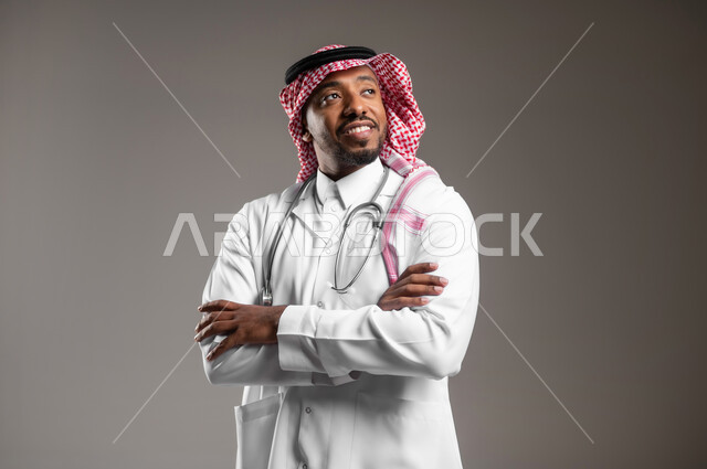 العمل في القطاع الصحي، النظر لمكان ما بايماءات الثقة بالنفس، مفهوم الفحص الطبي، تقديم الخدمات العلاجية للمرضى, بورتريه مقرب لطبيب عربي خليجي سعودي يرتدي الزي التقليدي والمعطف الطبي يضع السماعة الطبية يقف بأيدي متقاطعة، خلفية رمادية