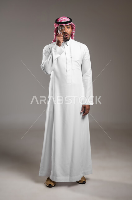 الفحص والاستكشاف والرؤية عن قرب، مفهوم توضيح الصورة، ايماءات السعادة والسرور، بورتريه لرجل عربي خليجي سعودي يرتدي الثوب التقليدي والشماغ ينظر من خلال العدسة المكبرة، التدقيق والتركيز باستخدام الأدوات البصرية، صورة لكامل الجسم، خلفية رمادية