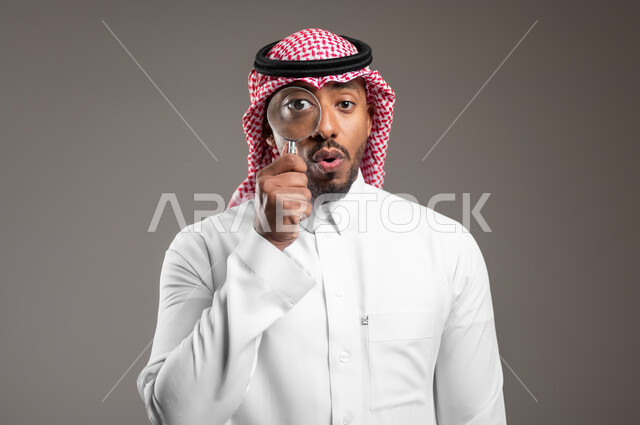 ايماءات السعادة والسرور، مفهوم توضيح الصورة، الفحص والاستكشاف والرؤية عن قرب، بورتريه مقرب لرجل عربي خليجي سعودي يرتدي الثوب التقليدي والشماغ ينظر من خلال العدسة المكبرة، التدقيق والتركيز باستخدام الأدوات البصرية، خلفية رمادية