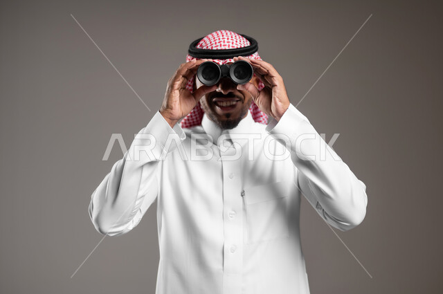 رؤية الفرص والتحديات المحتملة، حب المغامرة والاستكشاف، التخطيط المستقبلي للأهداف والطموحات، بورتريه مقرب لشاب عربي خليجي سعودي يرتدي الثوب التقليدي والشماغ ينظر من خلال المنظار ثنائي العينين، رؤية الاشياء البعيدة عبر التلسكوب، خلفية رمادية