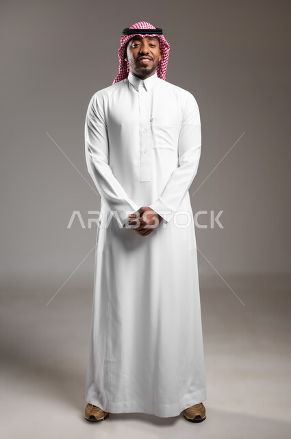 النظر إلى الكاميرا بايماءات الثقة بالنفس، تعابير الفرح والسعادة، مفهوم الاناقة والرجولة، الإهتمام بالمظهر الخارجي، بورتريه لرجل عربي خليجي سعودي يرتدي الثوب التقليدي والشماغ يقف بايدي متشابكة، صورة لكامل الجسم، خلفية رمادية