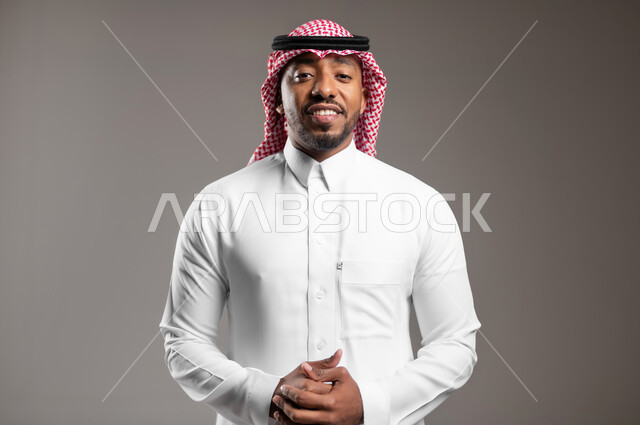 The concept of elegance and masculinity, looking at the camera with gestures of self-confidence, expressions of joy and happiness, interest in external appearance, close-up portrait of a Saudi Arabian Gulf man wearing a traditional thobe and shemagh standing with intertwined hands, gray background
