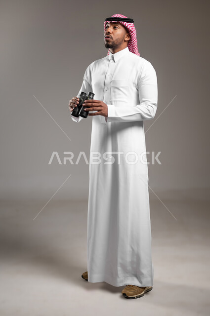 Seeing distant objects through a telescope, planning for future goals and ambitions, love of adventure and exploration, side portrait of a young Saudi Arabian Gulf man wearing traditional thobe and shemagh holding binoculars with shocked gestures, full body image, gray background