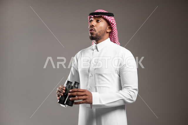 التخطيط المستقبلي للأهداف والطموحات، رؤية الفرص والتحديات المحتملة، حب المغامرة والاستكشاف، بورتريه مقرب من الجانب لشاب عربي خليجي سعودي يرتدي الثوب التقليدي والشماغ يحمل بيده منظار ثنائي العينين بايماءات الصدمة، رؤية الاشياء البعيدة عبر التلسكوب، خلفية رمادية