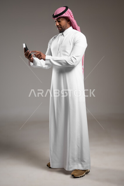 تطور التكنولوجيا والأجهزة التقنية، التسوق الالكتروني وتصفح برامج وتطبيقات الجوال، بورتريه من الجانب لرجل عربي خليجي سعودي يرتدي الثوب التقليدي والشماغ يحمل بيده الهاتف المحمول، الدردشة مع الأهل والأصدقاء، إيماءات الاندماج والتركيز، صورة لكامل الجسم، خلفية رمادية