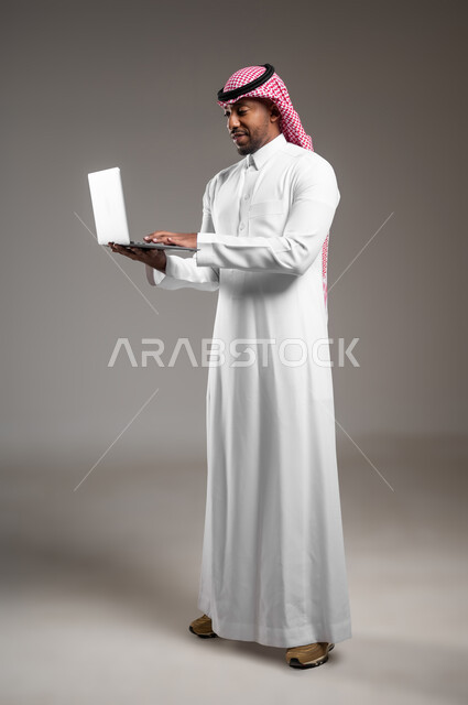 إنجاز مهام العمل من خلال اللابتوب، بورتريه من الجانب لرجل عربي خليجي سعودي يرتدي الثوب التقليدي والشماغ يحمل بيده جهاز الحاسوب المحمول، تصفح الإنترنت والعمل عن بعد، الضغط على أزرار لوحة المفاتيح، صورة لكامل الجسم، خلفية رمادية