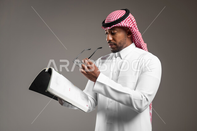 قضاء وقت الفراغ بالتعلم والثقافة، ايماءات الاندماج والتركيز، مفهوم التثقيف والبحث العلمي، بورتريه مقرب لشاب عربي خليجي سعودي مبتسم يرتدي الثوب التقليدي والشماغ يحمل بيده النظارة الطبية يقوم بقراءة كتاب, هواية المطالعة والاستكشاف، خلفية رمادية