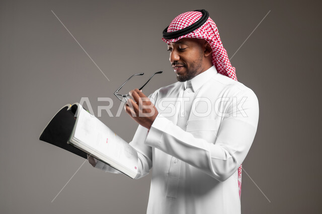 مفهوم التثقيف والبحث العلمي، قضاء وقت الفراغ بالتعلم والثقافة، ايماءات الاندماج والتركيز، بورتريه مقرب لشاب عربي خليجي سعودي مبتسم يرتدي الثوب التقليدي والشماغ يحمل بيده النظارة الطبية يقوم بقراءة كتاب, هواية المطالعة والاستكشاف، خلفية رمادية