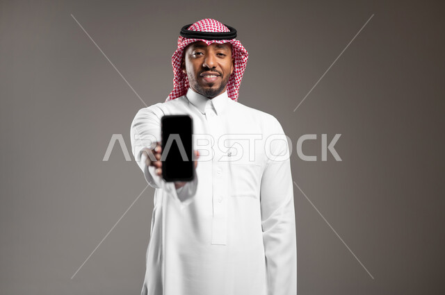 Using modern technology, displaying a blank black screen, quality mobile applications and services, close-up portrait of a smiling Saudi Arabian Gulf man wearing traditional thobe and shemagh looking at the camera with gestures of pleasure, gray background