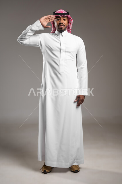 الإحترام وإطاعة الأوامر، مفهوم التحية والسلام، بورتريه لرجل عربي خليجي سعودي يرتدي الثوب التقليدي والشماغ يرفع يده اليمنى لأداء التحية العسكرية، صوره الجسم بالطول الكامل، خلفيه رماديه