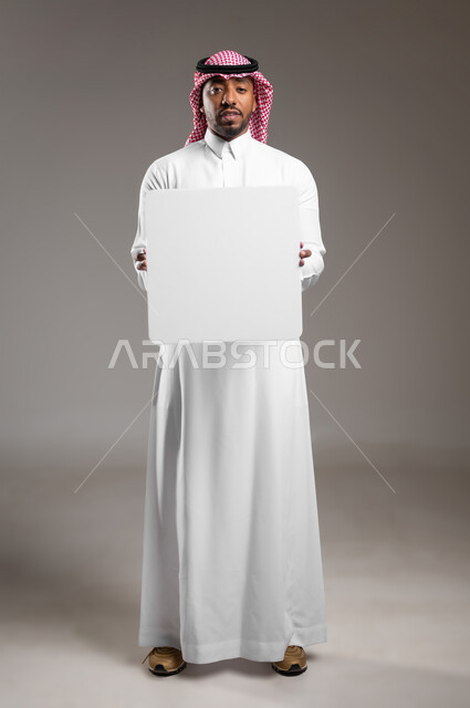 لوح إعلاني فارغ، موك اب مستطيل أبيض، بورتريه لرجل عربي خليجي سعودي يرتدي الثوب التقليدي والشماغ يحمل بيده لوحه بيضاء، الوقوف باستقامه والنظر إلى الكاميرا بايماءات الثقة بالنفس، صورة الجسم بالطول الكامل، خلفية رماديه