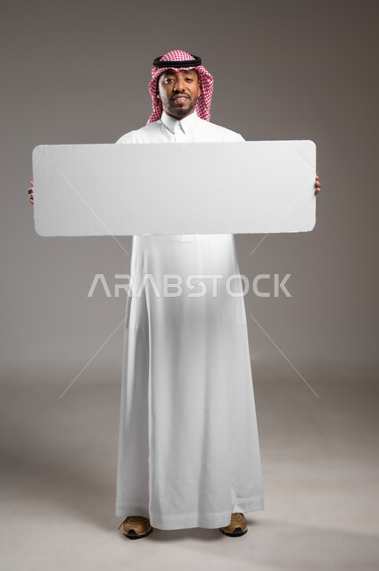 لوح إعلاني فارغ، موك اب مستطيل أبيض، بورتريه لرجل عربي خليجي سعودي يرتدي الثوب التقليدي والشماغ يحمل بيده لوحه بيضاء، الوقوف باستقامه والنظر إلى الكاميرا بايماءات الثقة بالنفس، صورة الجسم بالطول الكامل، خلفية رماديه