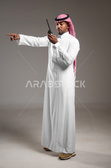 الإستجابة لحالات الطوارئ، المتابعة والإشراف على خطط العمل، بورتريه من الجانب لرجل عربي خليجي سعودي مبتسم يرتدي الثوب التقليدي والشماغ يقوم باستخدم جهاز الارسال والاستقبال اللاسلكي، مفهوم ادارة المشاريع عن بعد، صورة لكامل الجسم، خلفية رمادية