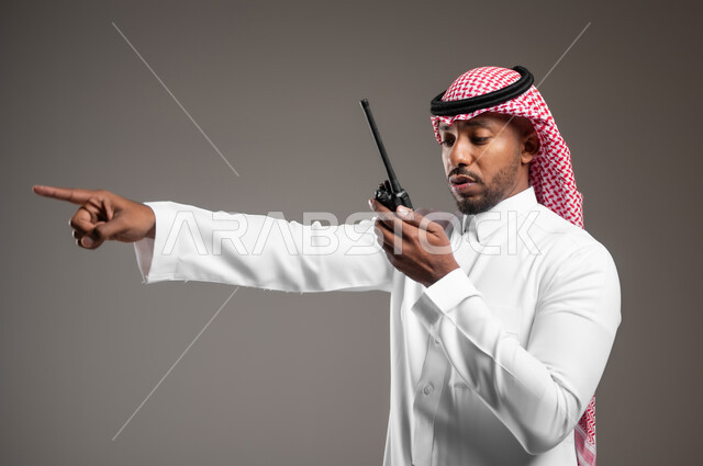 مفهوم ادارة المشاريع عن بعد، الإستجابة لحالات الطوارئ، المتابعة والإشراف على خطط العمل، بورتريه مقرب من الجانب لرجل عربي خليجي سعودي مبتسم يرتدي الثوب التقليدي والشماغ يقوم باستخدم جهاز الارسال والاستقبال اللاسلكي، خلفية رمادية