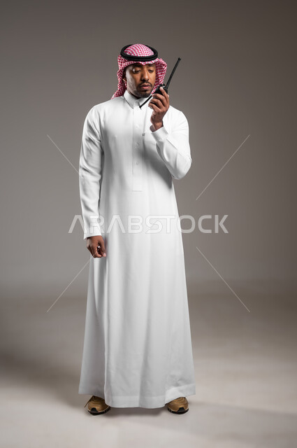 المتابعة والإشراف على خطط العمل، الإستجابة لحالات الطوارئ، بورتريه لرجل عربي خليجي سعودي مبتسم يرتدي الثوب التقليدي والشماغ يقوم باستخدم جهاز الارسال والاستقبال اللاسلكي، مفهوم ادارة المشاريع عن بعد، صورة لكامل الجسم، خلفية رمادية