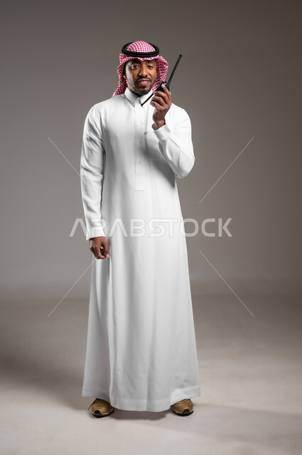 الإستجابة لحالات الطوارئ، المتابعة والإشراف على خطط العمل، بورتريه لرجل عربي خليجي سعودي مبتسم يرتدي الثوب التقليدي والشماغ يقوم باستخدم جهاز الارسال والاستقبال اللاسلكي، مفهوم ادارة المشاريع عن بعد، صورة لكامل الجسم، خلفية رمادية