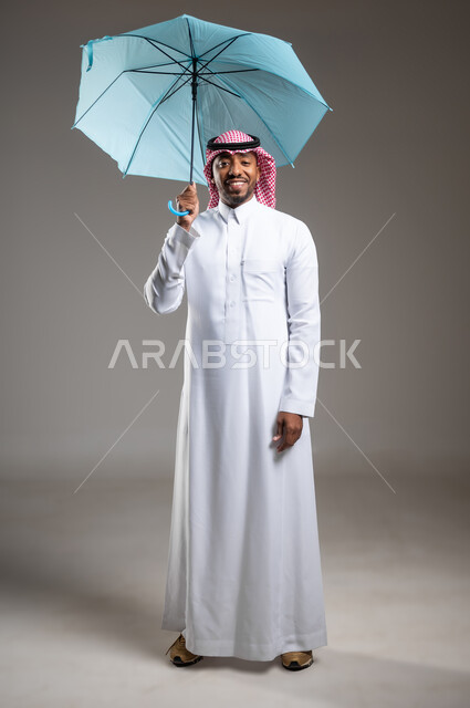 الحماية من الأمطار في الشتاء، تعابير البهجة والسرور، بورتريه لشاب عربي خليجي سعودي مبتسم يرتدي الثوب التقليدي والشماغ يحمل بيده مظلة زرقاء، ايماءات الفرح والسعادة، التظلل من أشعة الشمس، صورة لكامل الجسم، خلفية رمادية