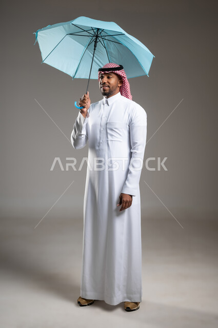 تعابير البهجة والسرور، بورتريه لشاب عربي خليجي سعودي مبتسم يرتدي الثوب التقليدي والشماغ يحمل بيده مظلة زرقاء، ايماءات الفرح والسعادة، التظلل من أشعة الشمس، الحماية من الأمطار في الشتاء، صورة لكامل الجسم، خلفية رمادية