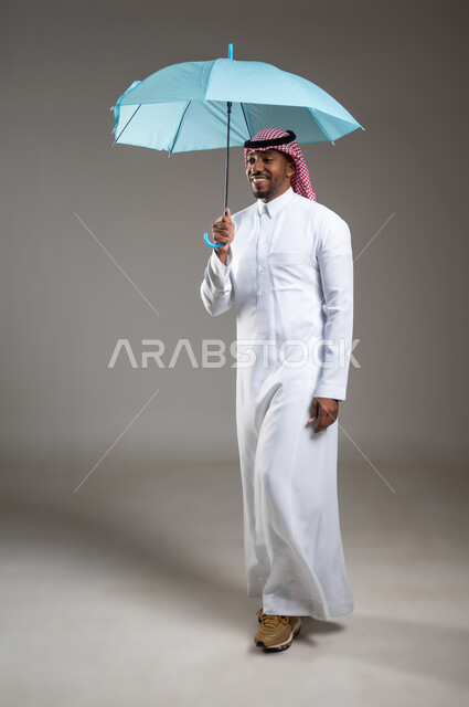 التظلل من أشعة الشمس، تعابير البهجة والسرور، بورتريه لشاب عربي خليجي سعودي مبتسم يرتدي الثوب التقليدي والشماغ يحمل بيده مظلة زرقاء، ايماءات الفرح والسعادة، الحماية من الأمطار في الشتاء، صورة لكامل الجسم، خلفية رمادية