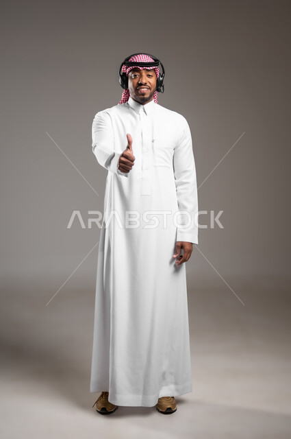 تعابير الموافقة والقبول، العمل في مجال خدمة العملاء والدعم الفني، الرد على استفسارات الزبائن، استخدام تقنية الهيدفون، بورتريه لرجل عربي خليجي سعودي يرتدي الثوب التقليدي والشماغ يضع سماعة الاتصال يرفع يده بايماءات الاعجاب، صورة لكامل الجسم، خلفية رمادية